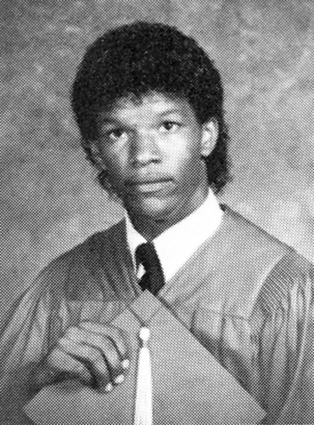 Black and white photo of a young person wearing a graduation gown and holding a tassel. They have short, curly hair and a serious expression, standing against a textured studio backdrop.
