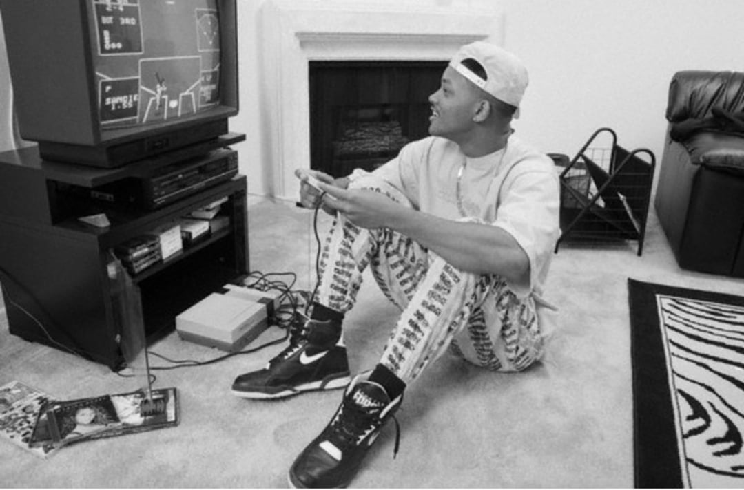 A young man wearing a cap backward, patterned pants, and Nike sneakers sits on the floor playing a video game on an old TV in a living room, smiling as he looks at the screen.