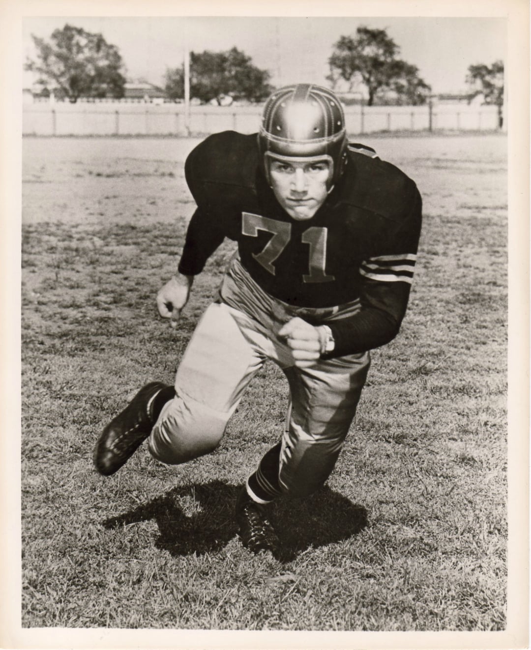 Black-and-white photo of a football player wearing a vintage uniform and helmet, number 71, running on a grass field with trees and a fence in the background.