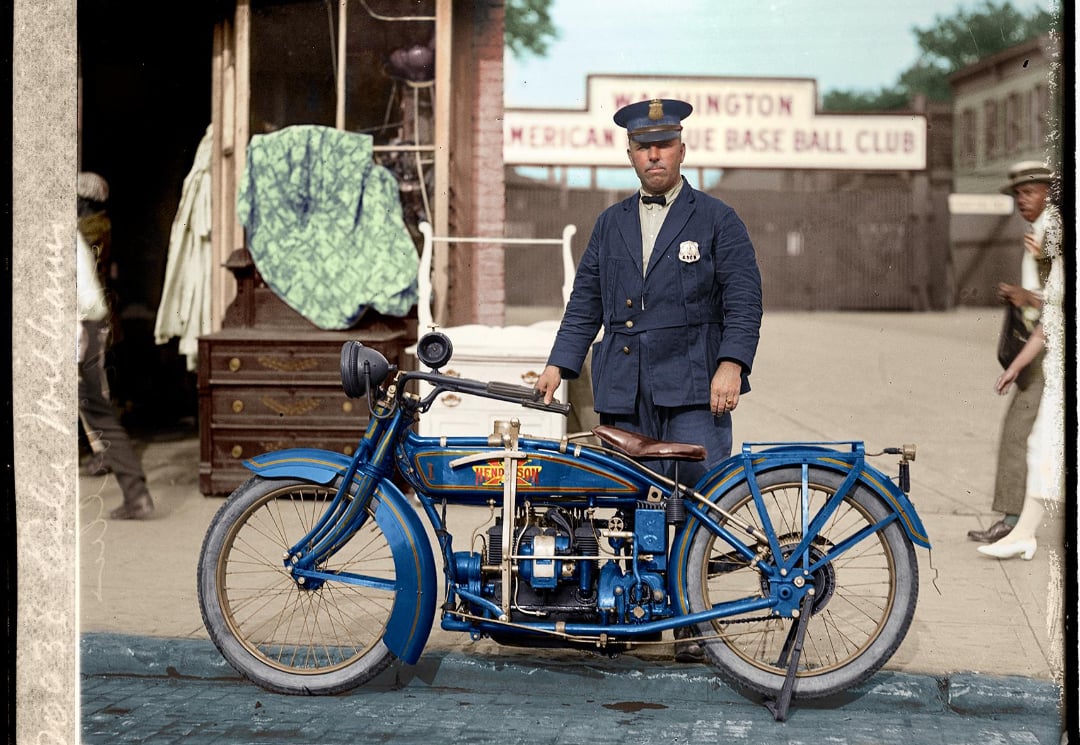 A police officer in uniform stands behind a vintage blue motorcycle on a cobblestone street, with a wooden dresser and a sign reading "American Base Ball Club" visible in the background.
