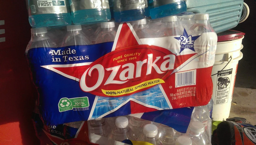 A 24-pack of Ozarka 100% natural spring water bottles with a red and blue label featuring a large white star and text reading "Made in Texas." The package is stacked with other bottled drinks.