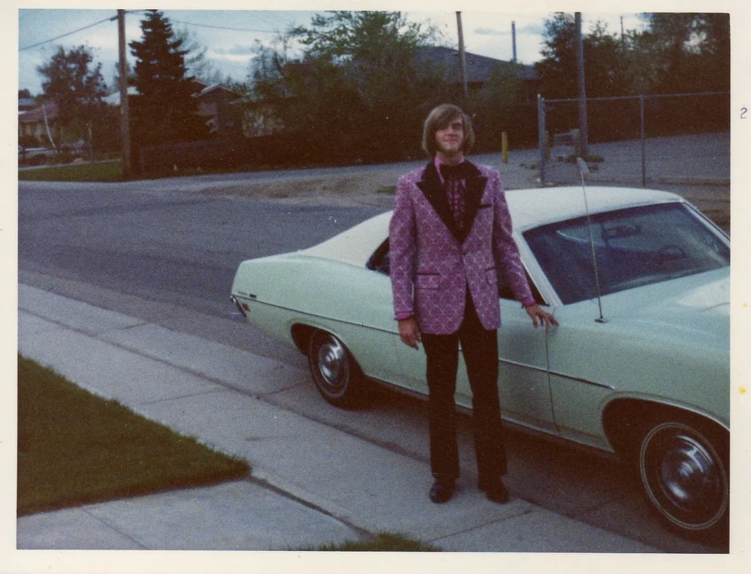 A person wearing a pink patterned blazer, dark pants, and a bow tie stands on a sidewalk next to a light green vintage car in a suburban neighborhood. Trees, houses, and power lines are visible in the background.