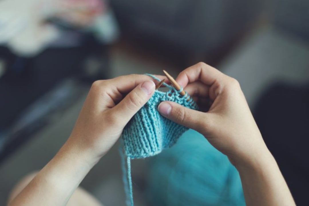 A person’s hands holding wooden knitting needles and working with light blue yarn, creating a knitted fabric. The background is blurred, focusing on the knitting process.