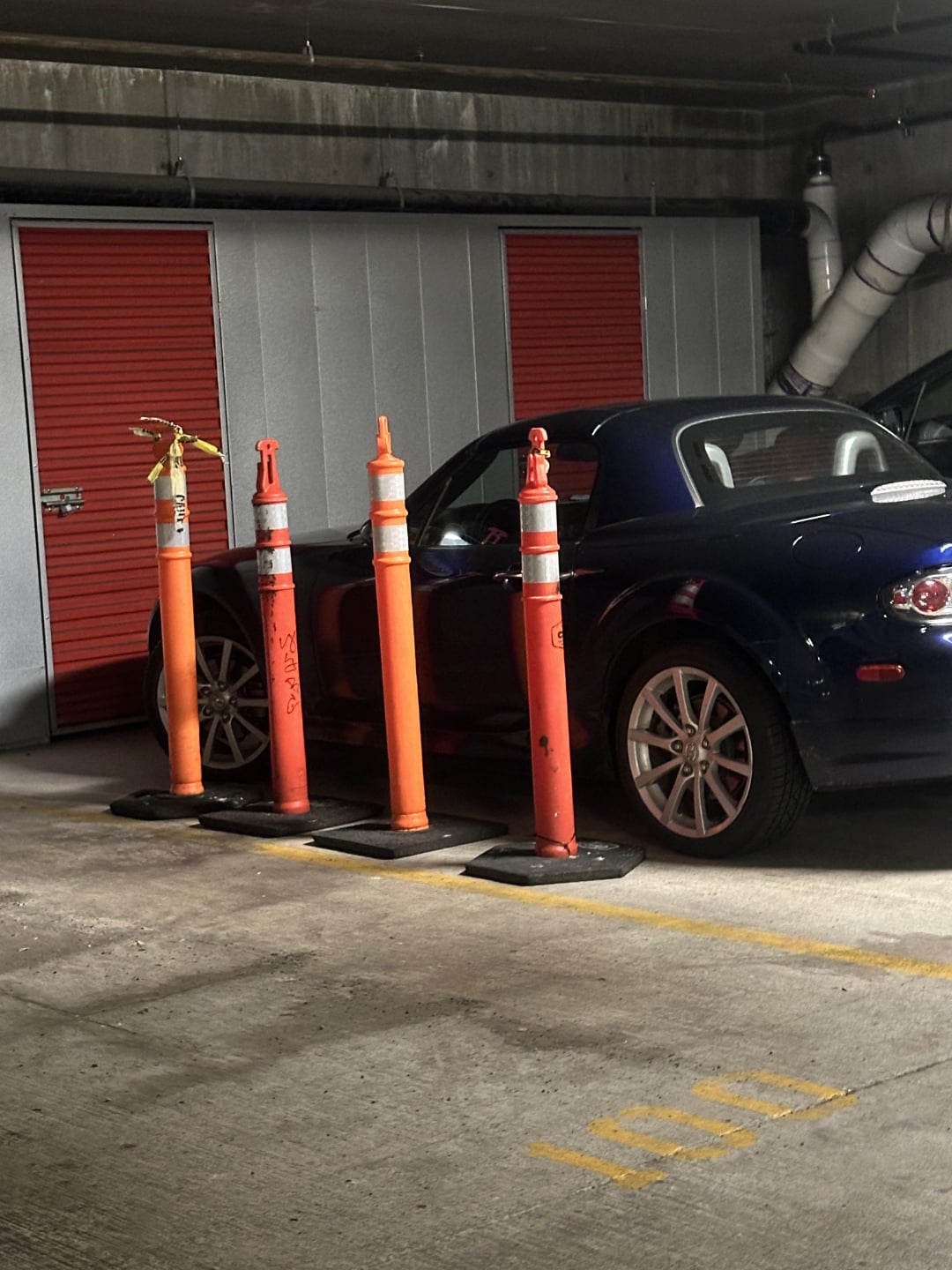 A dark blue convertible car is parked in a garage space, partially blocked by four orange traffic cones standing in front of it. There are red storage doors in the background.