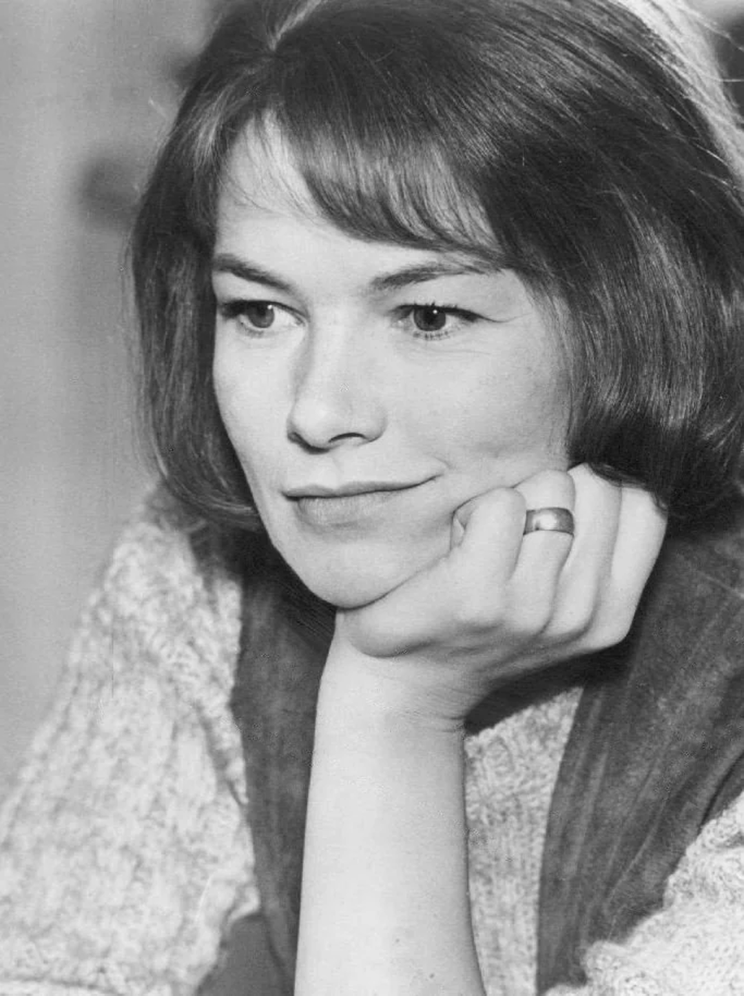 A woman with short, straight hair rests her chin on her hand, wearing a thoughtful expression. She is dressed in a light-colored sweater and wears a ring on her finger. The image is black and white.