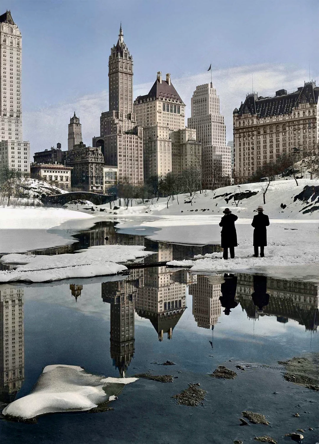 Two people in dark coats stand by a snow-covered, partially frozen pond, with tall city buildings reflected in the water. The scene has a wintery, early 20th-century urban atmosphere.