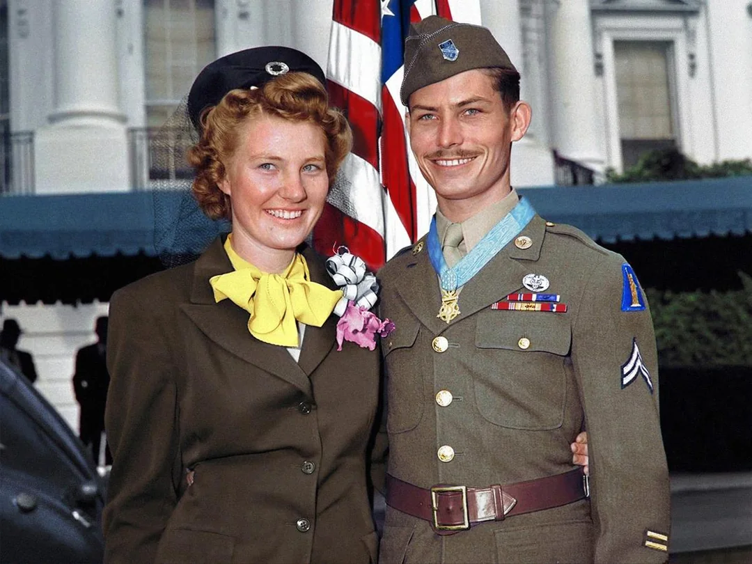A smiling woman and man in military uniforms stand together in front of an American flag and a building, likely a government building. The man wears medals on his uniform and they both appear happy.