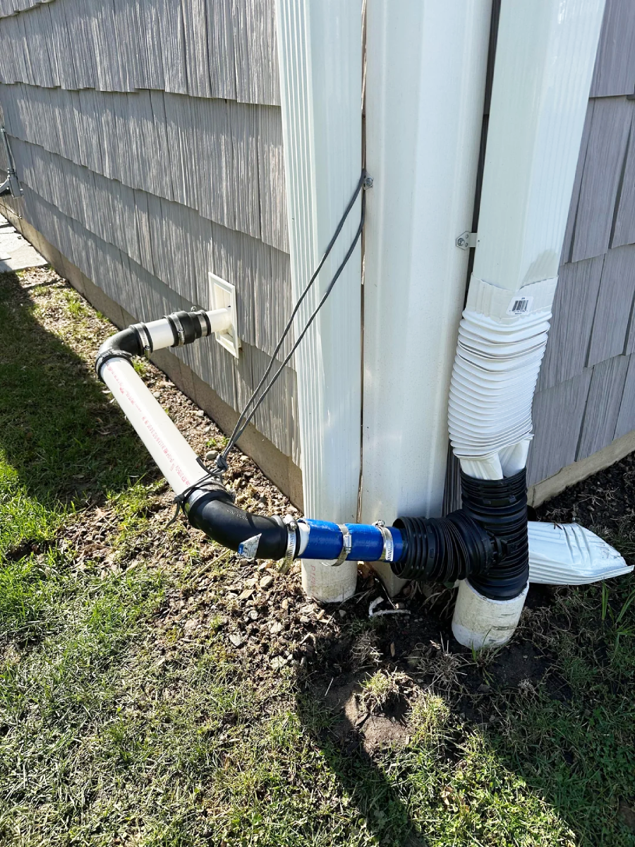 A close-up of a house’s exterior showing an elaborate gutter and drainage setup with multiple pipes, connectors, and hoses directing water away from the foundation onto the lawn.