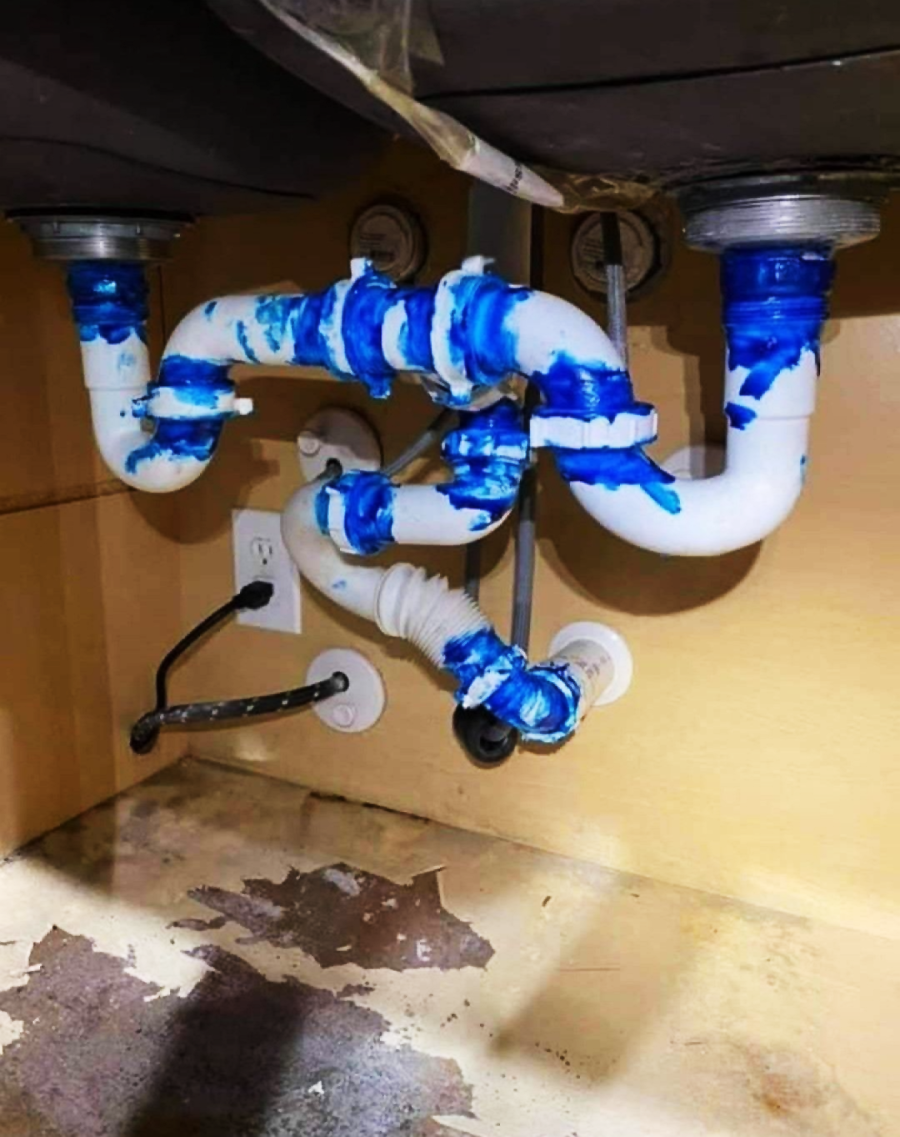 White PVC pipes under a sink with excessive blue sealant applied at all joints and connections; the pipes are awkwardly arranged, and the cabinet floor shows water damage.