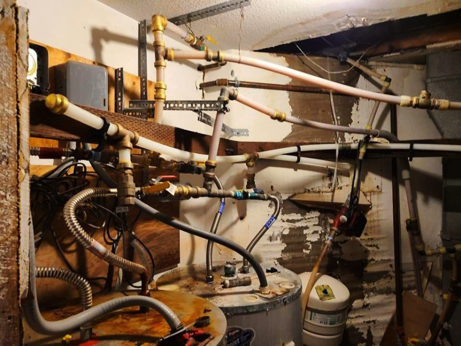 A cluttered utility room with exposed pipes, wires, and valves connected to a water heater. The walls show signs of water damage and peeling paint, and various plumbing parts are scattered around.
