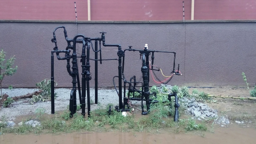A cluster of black metal pipes and valves stands on wet ground with some puddles and sparse grass, in front of a brown wall with a red section at the top.