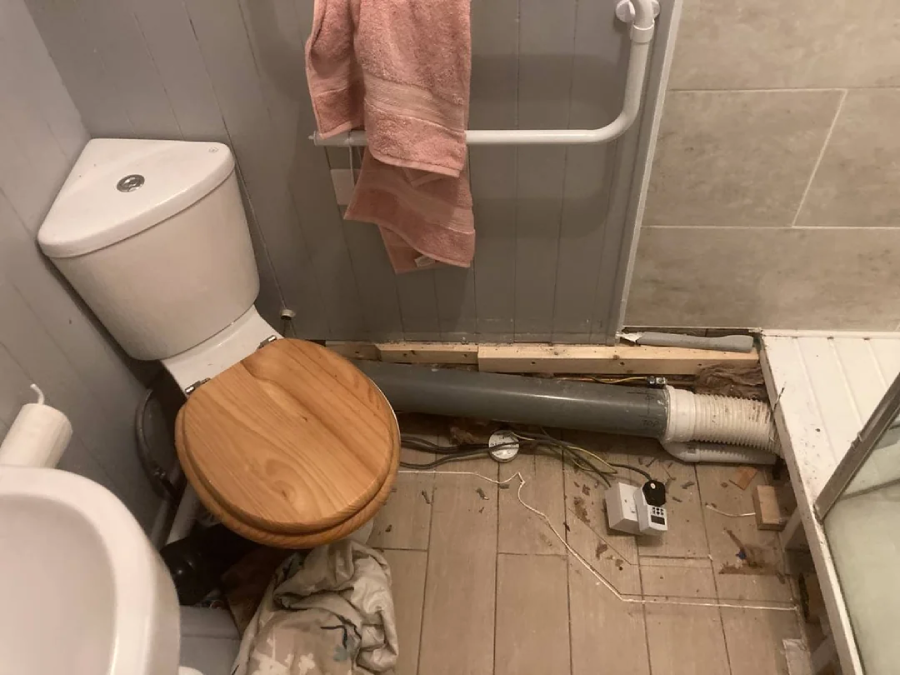 A bathroom with a toilet beside a shower; a large gray pipe runs along the floor in front of the toilet and into the shower. The area is messy with debris and exposed wires. A pink towel hangs on a rail.