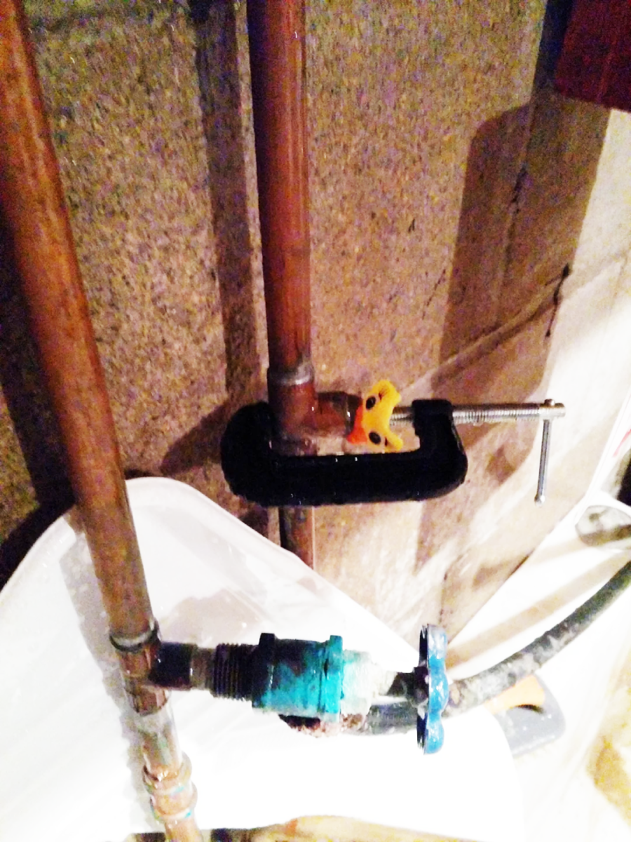 A clamp holding two copper pipes together against a concrete wall, with a yellow valve and a blue water pipe connector visible below. Some water stains and corrosion are present on the pipes.