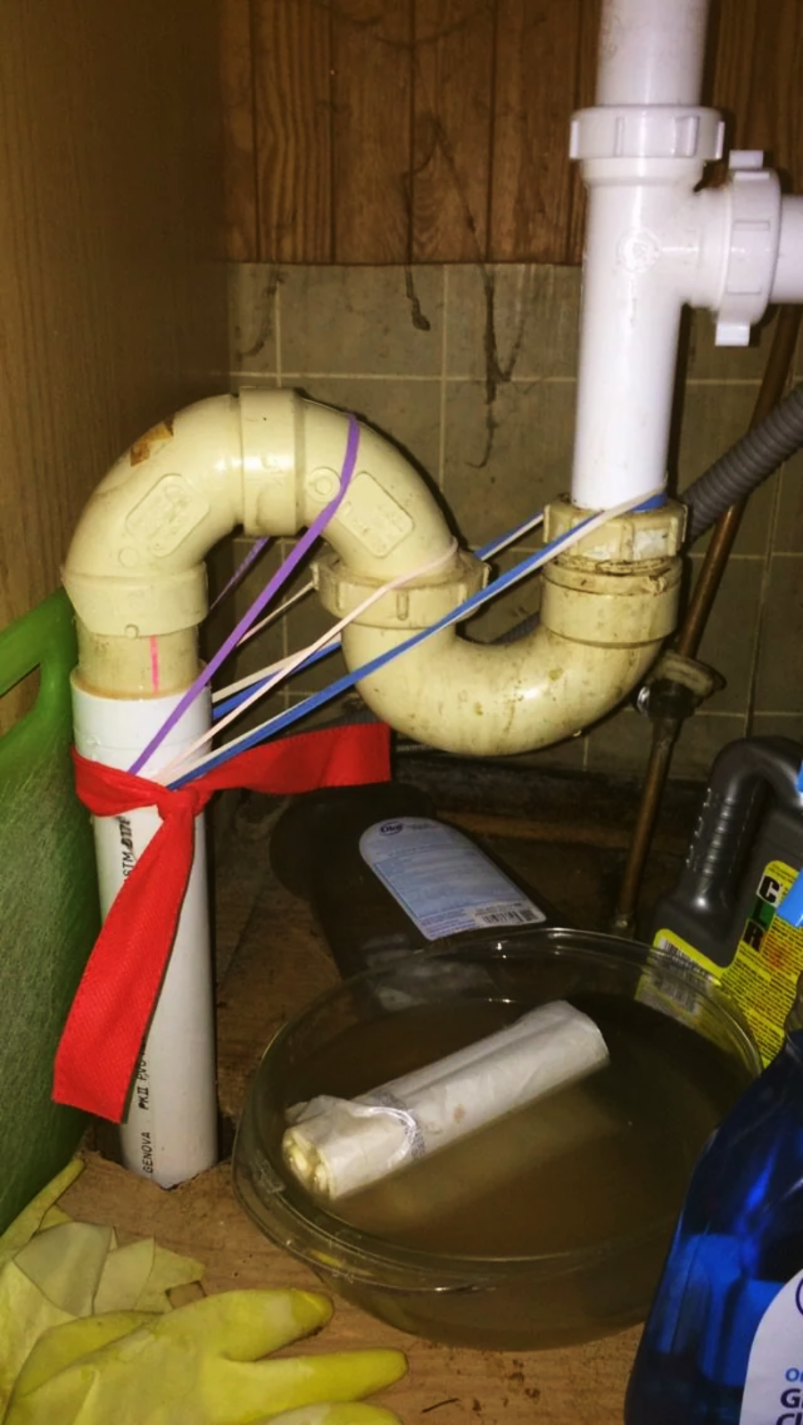 A leaking kitchen drain pipe is held together with zip ties, rubber bands, and a red cloth, above a bowl catching water. Cleaning supplies and gloves are nearby.