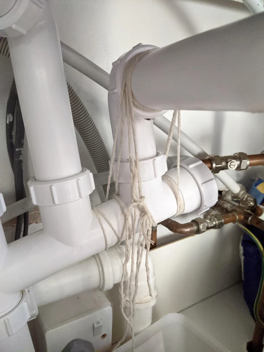 White plumbing pipes under a sink are loosely tied together with string, with several strands hanging down. Copper pipes and part of a blue object are visible in the background.