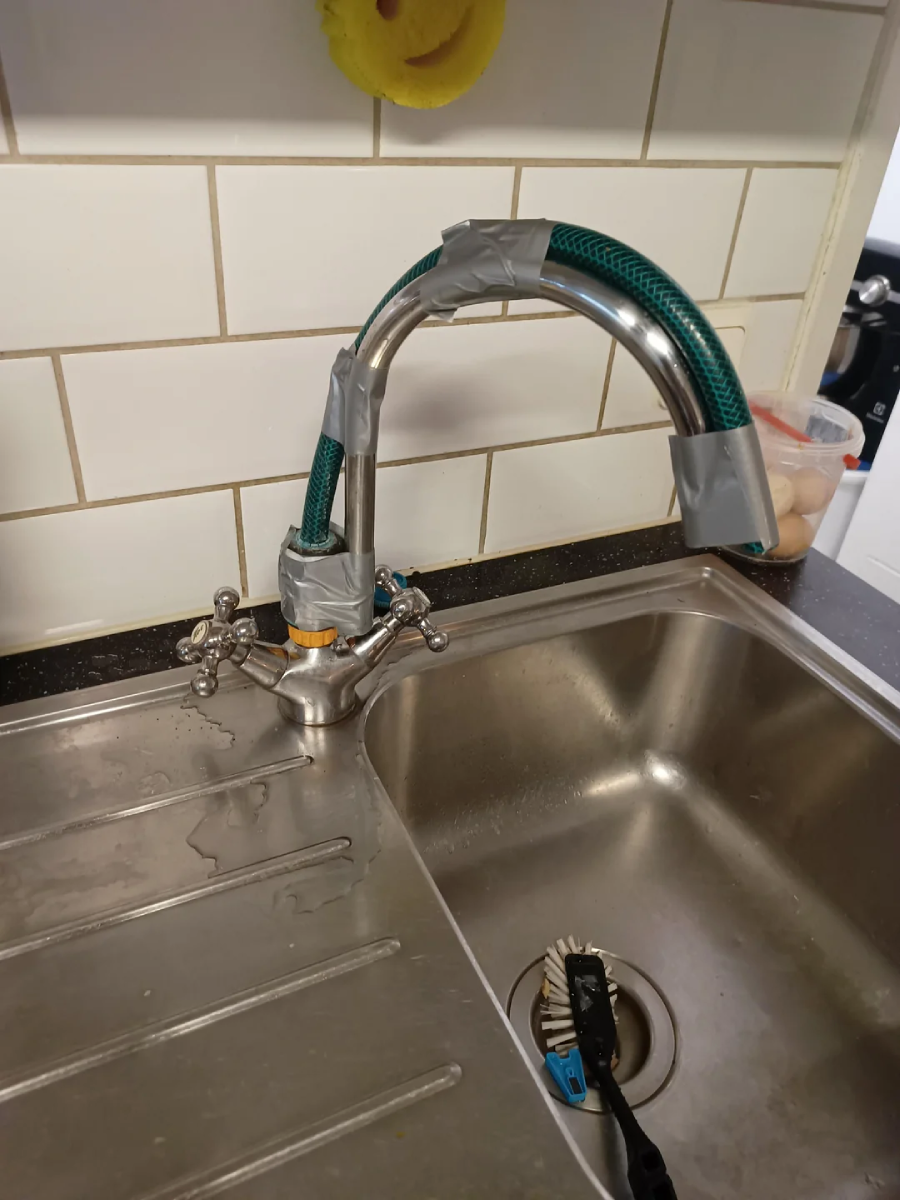 A kitchen faucet is repaired with a green garden hose and duct tape, with the hose attached to the spout and base. The sink is stainless steel, and a scrub brush and a yellow sponge are visible nearby.