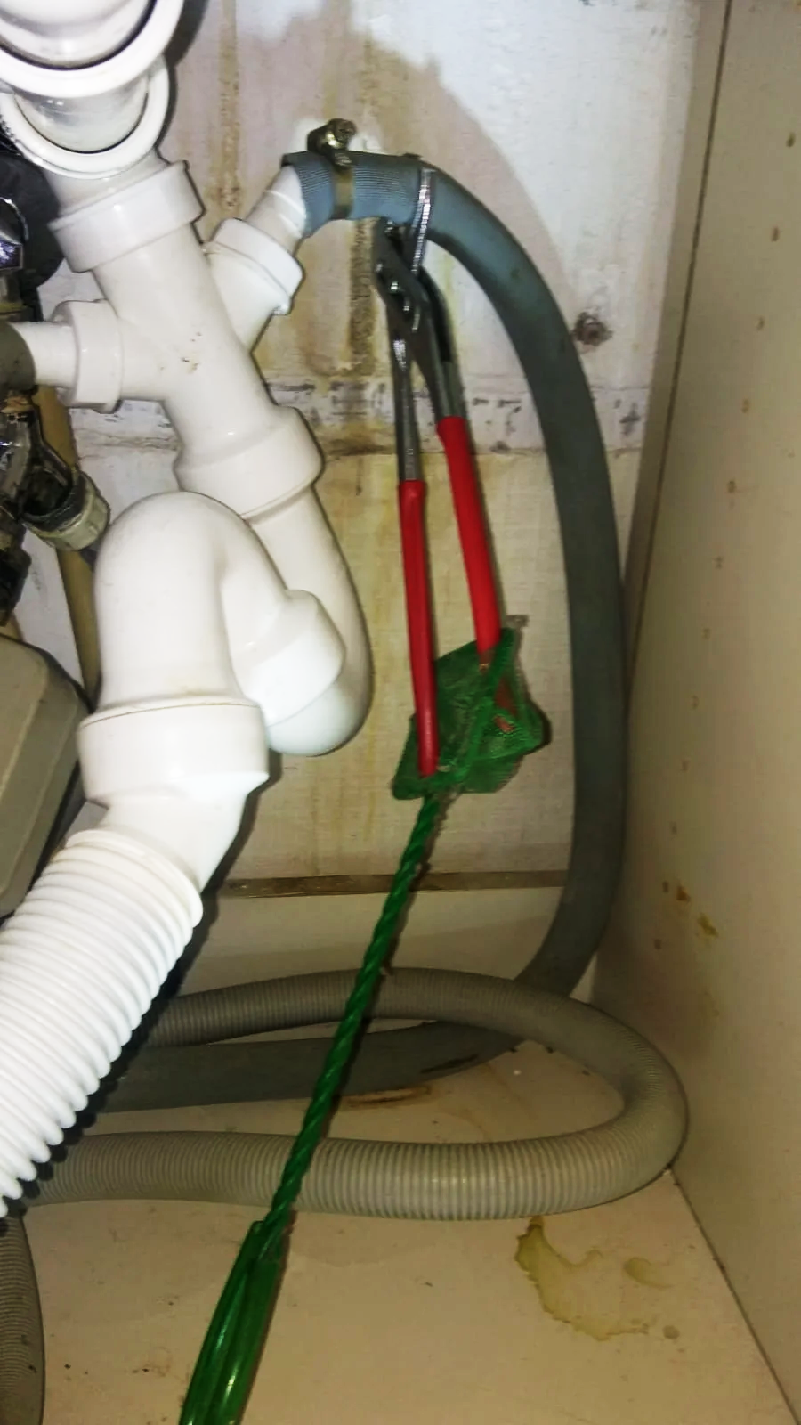 A pair of red-handled pliers is clamped onto a grey pipe under a sink, with green string tied to the pliers and pulling downward. White plastic plumbing pipes and hoses are also visible nearby.