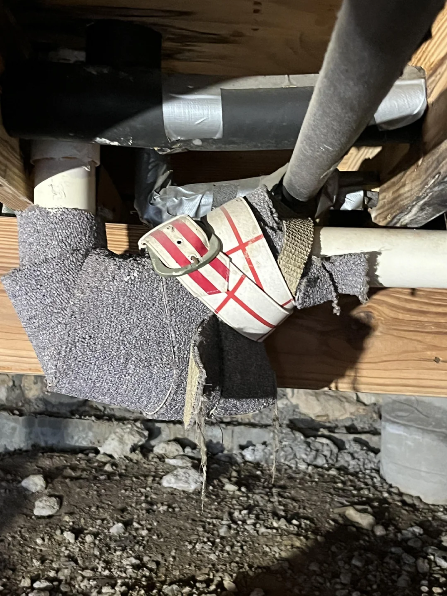 A close-up of improvised pipe insulation and repairs under a wooden structure, showing fabric, tape, and zip ties wrapped around PVC pipes above a dirt floor.