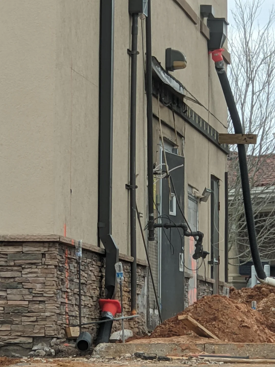 A construction site with dirt piles and pipes attached to the side of a beige building with stone accents. Some pipes are taped with red material and lead from the ground up to the roof.