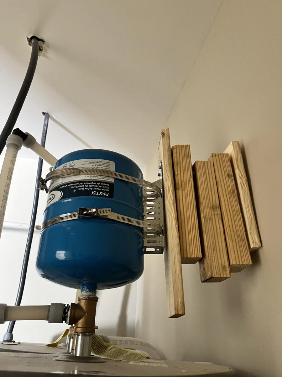 A blue pressure tank is mounted on a wall with metal straps and brackets. Next to it, several uneven wooden planks are attached vertically to the wall, possibly as spacers or supports. Pipes are visible nearby.