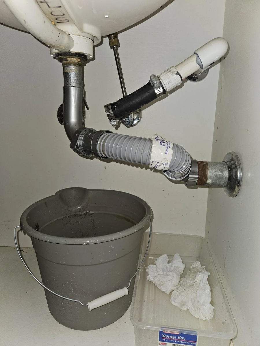 A grey plastic bucket is placed under a sink drainpipe, which has makeshift repairs with tape and a flexible hose. Nearby are a clear plastic box and some crumpled paper towels.