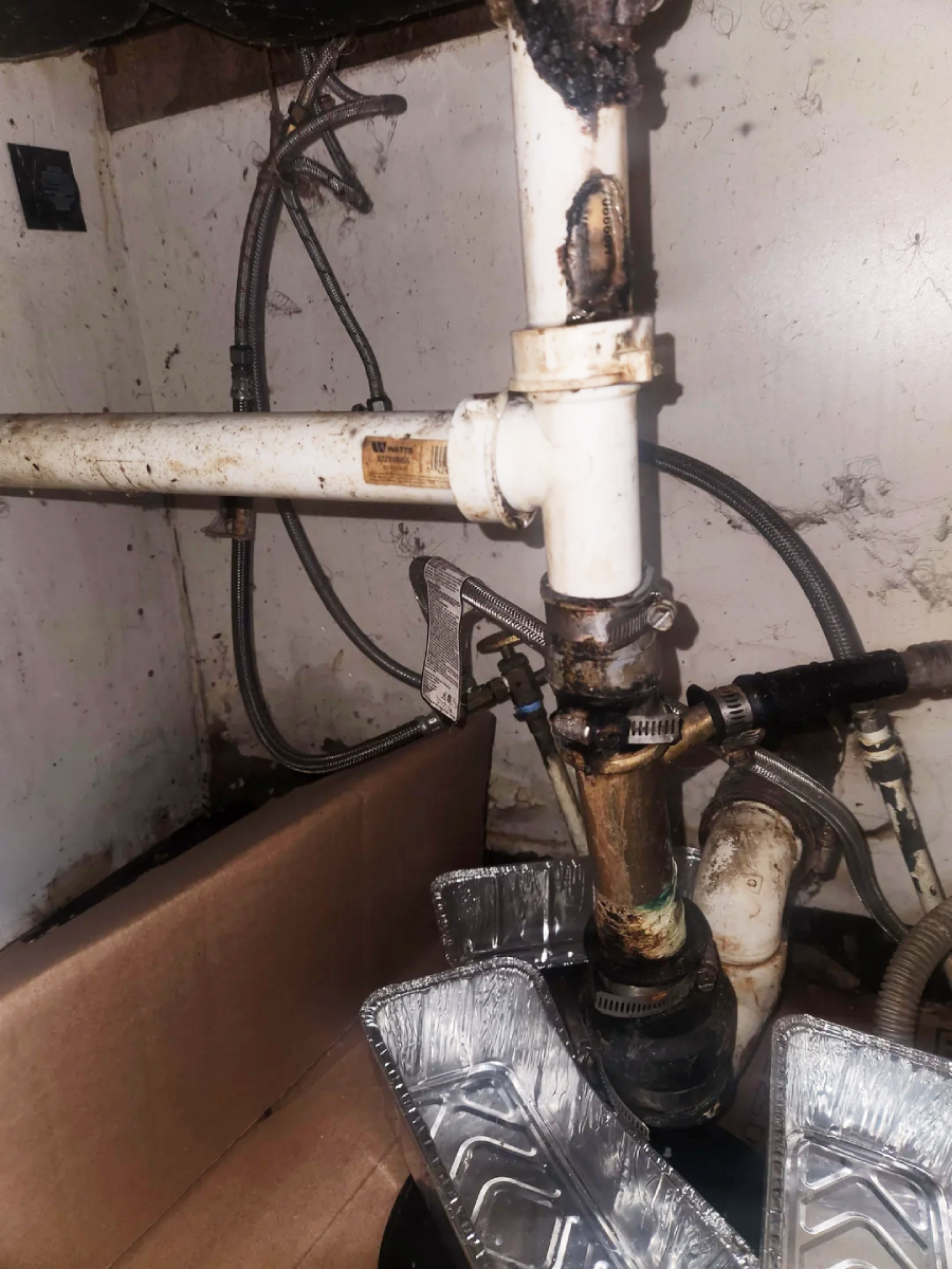 Plumbing pipes under a sink with visible rust, grime, and leaks. Aluminum trays are placed underneath to catch dripping water. The wall behind is stained, indicating water damage or mold.