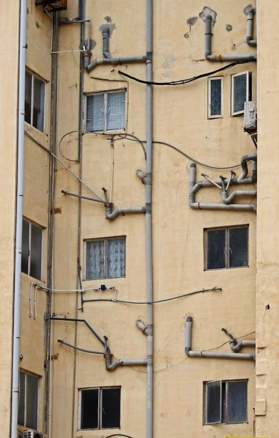 A beige apartment building wall with multiple small windows and numerous gray pipes and cables running haphazardly in various directions across the surface.