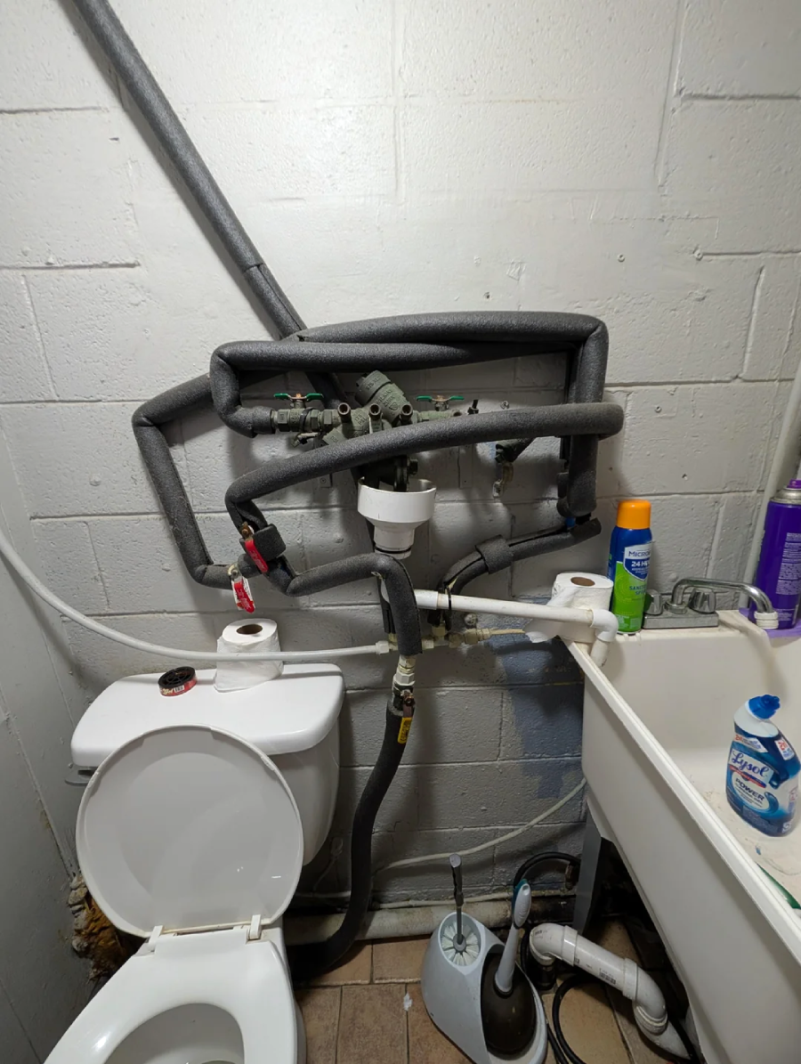 A small bathroom with exposed insulated pipes and valves running above a white toilet and next to a utility sink. Cleaning supplies are on the sink, and a toilet brush is on the floor nearby.