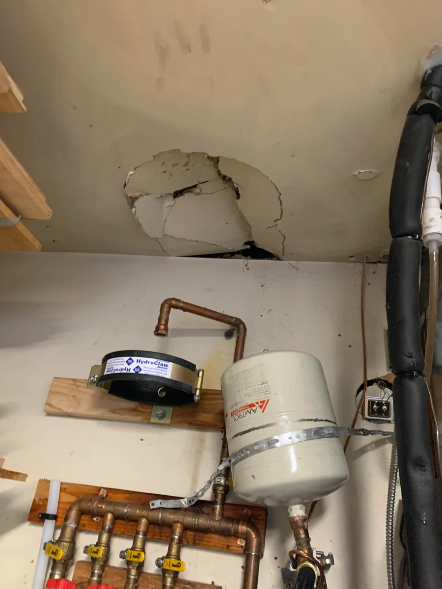 A ceiling with a large, cracked hole and water damage above exposed pipes and two water tanks mounted on a wall in a utility or boiler room.