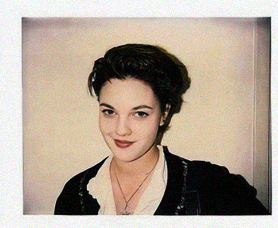 A young person with short dark hair and a slight smile poses in front of a plain light-colored wall, wearing a black jacket over a white-collared top and a necklace.
