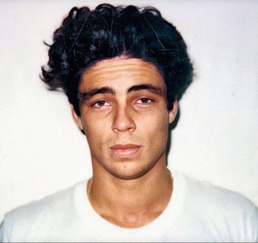 A young man with wavy dark hair, wearing a white shirt, poses against a plain white background, looking directly at the camera with a neutral expression.