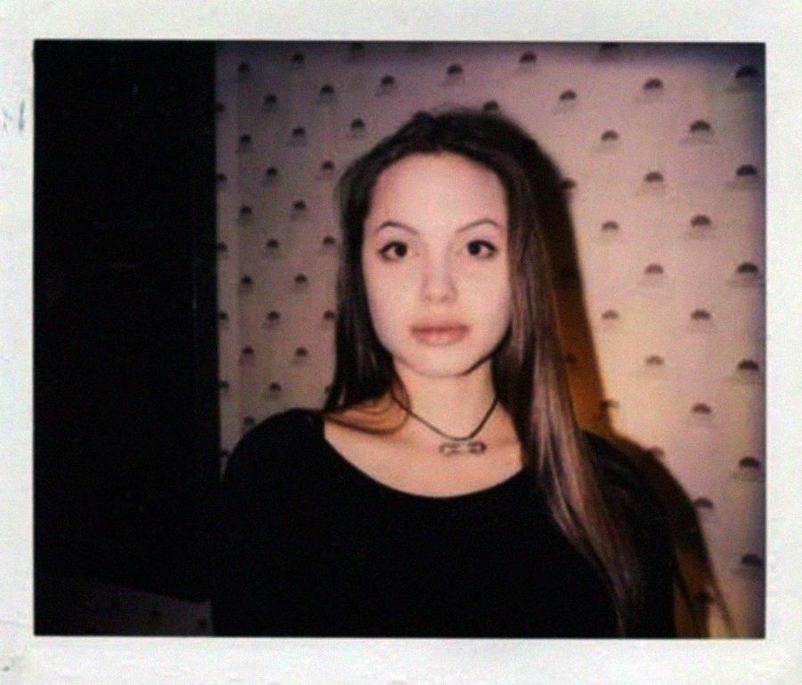 A young woman with long brown hair and a black top stands in front of a light-colored, patterned wall, looking directly at the camera in a polaroid-style photograph.