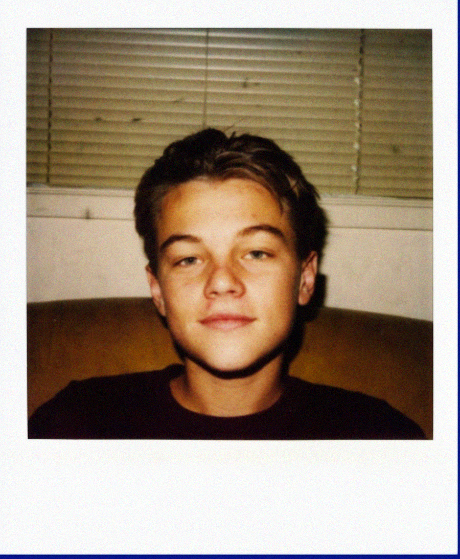 A young person with short brown hair sits on a couch, smiling slightly. The background shows closed blinds on a window. The image has a Polaroid-style white border.