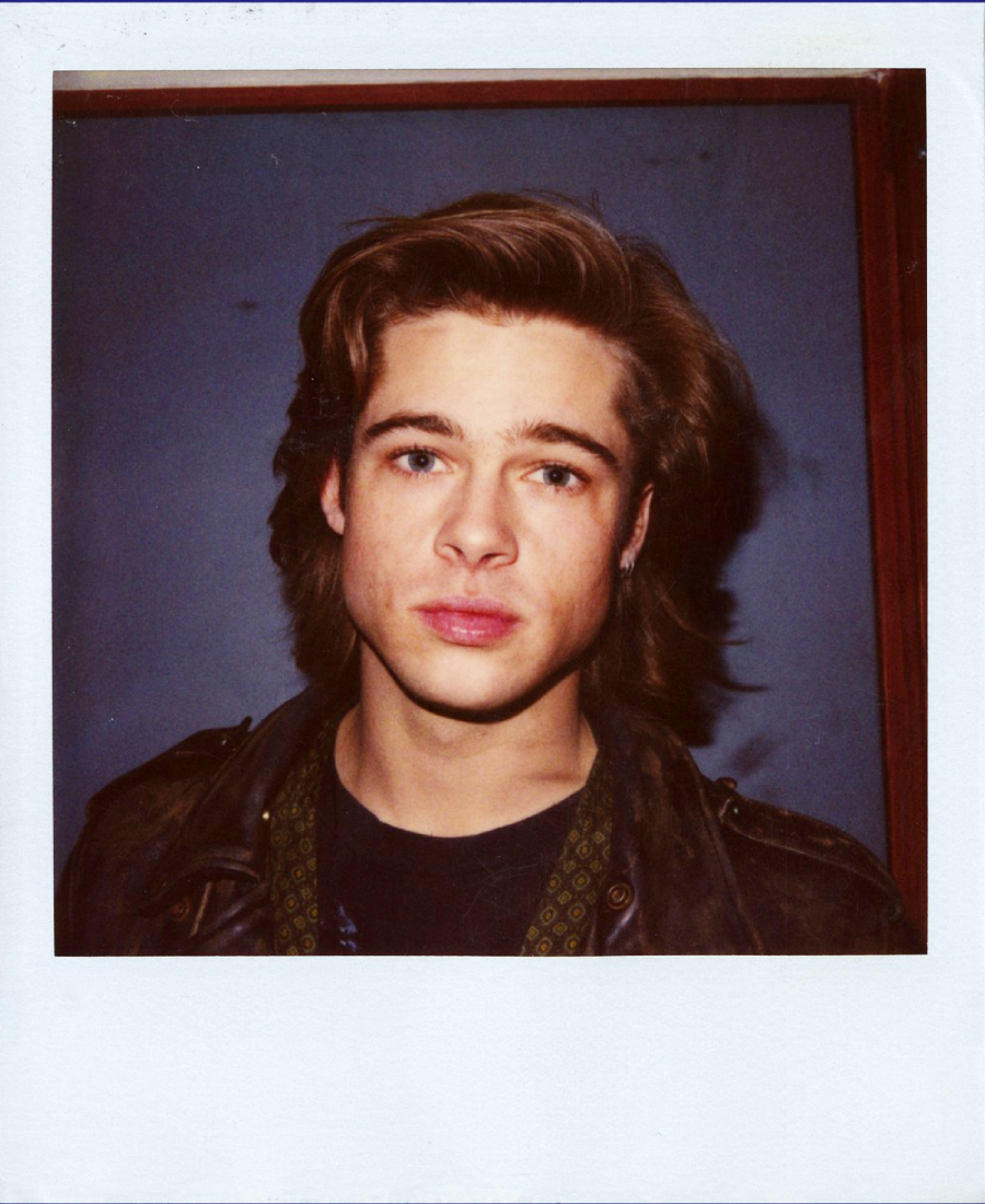 A young man with medium-length light brown hair stands against a plain blue background, wearing a dark leather jacket and patterned scarf, looking directly at the camera with a neutral expression.