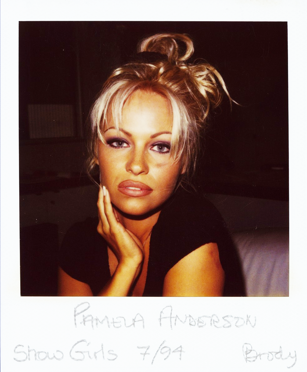A woman with blonde hair styled in a messy updo poses with her hand on her face, wearing a black top and makeup. The photo has a Polaroid frame labeled “Pamela Anderson Show Girls 7/94 Brody.”