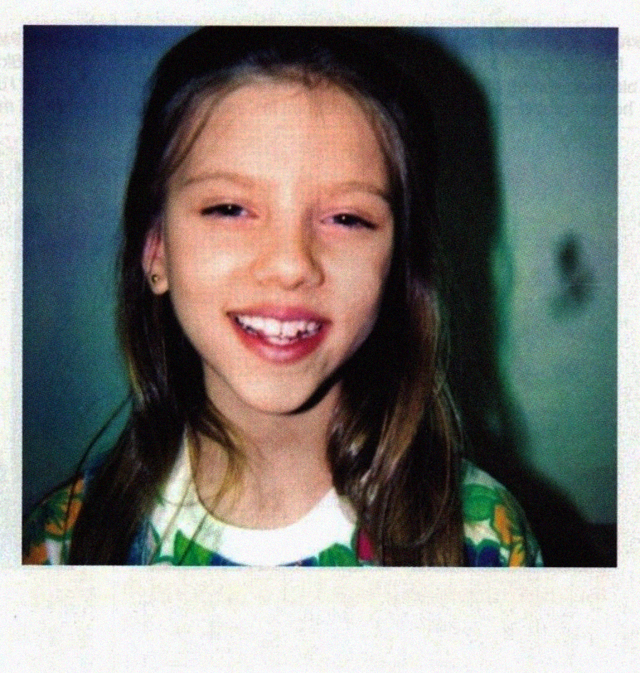 A young girl with long brown hair smiles at the camera. She is wearing a colorful patterned shirt and has a missing front tooth. The background is plain and softly lit.