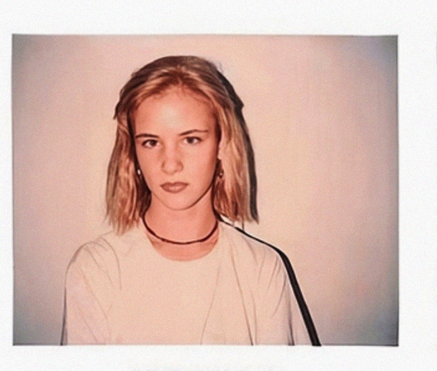 A young woman with shoulder-length blonde hair wearing a white shirt stands against a plain light-colored background, looking directly at the camera with a neutral expression.
