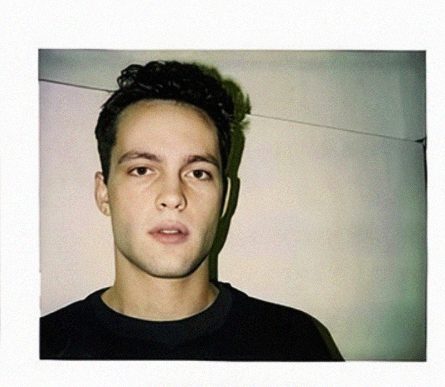 A young man with short, dark hair and a neutral expression stands against a plain, light-colored wall, wearing a dark shirt. The lighting casts a soft shadow on the wall behind him.