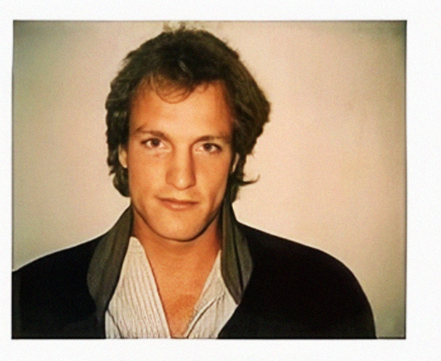 A young man with light brown hair and a slight smile faces the camera. He is wearing a dark jacket over a light-colored, partially unbuttoned shirt, standing against a plain off-white background.