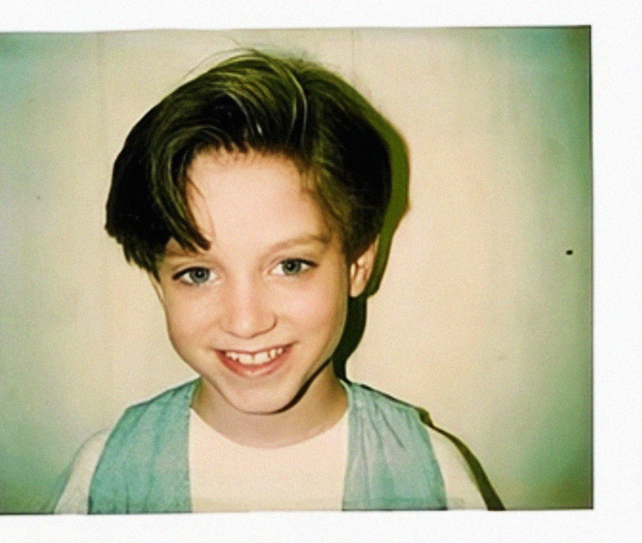 A smiling young child with light skin, blue eyes, and short brown hair poses in front of a plain background. The child is wearing a white shirt and a light blue vest. The image has a vintage, Polaroid-style border.