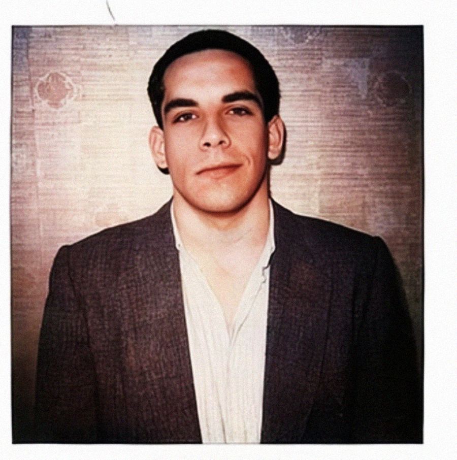 A young man with short dark hair, wearing a blazer over a light-colored shirt, stands against a textured wall and looks calmly at the camera in a Polaroid-style photo.