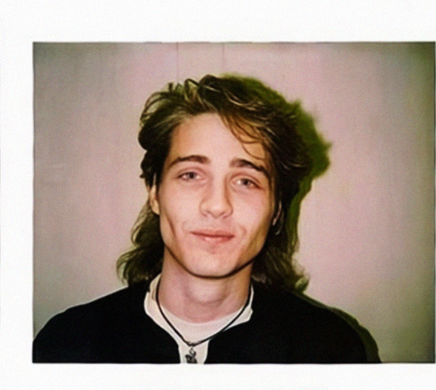 A young person with long, wavy hair and a slight smile poses for a Polaroid-style portrait against a plain background, wearing a dark shirt and a necklace.