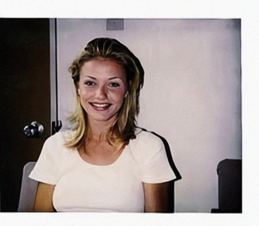 A young woman with blonde hair, wearing a white short-sleeved top, sits and smiles at the camera. The background includes a door and part of a chair.
