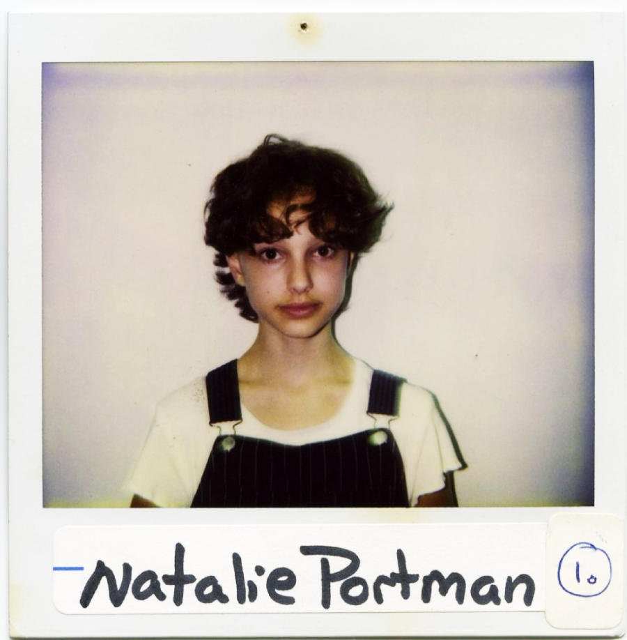 A young person with short, curly hair wears a white shirt and dark overalls, standing against a plain light background. The photo is in Polaroid style with “Natalie Portman” handwritten at the bottom.