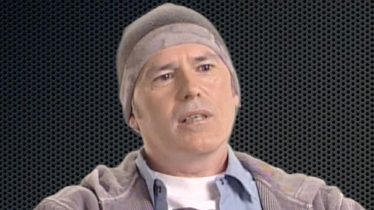 A person wearing a gray beanie, light blue shirt, and gray jacket sits in front of a black, dotted background, looking slightly to the side with a neutral expression.