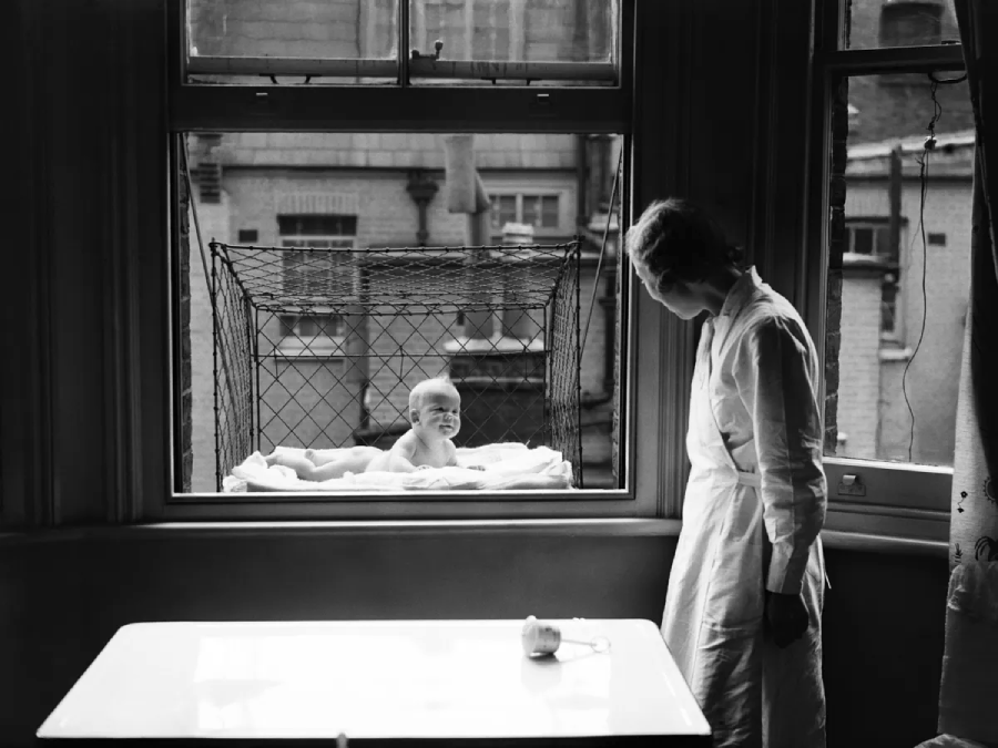 A woman stands by a window, looking at a baby lying in a wire mesh crib suspended outside the window of an apartment building. The baby is lying on a blanket, with city buildings visible in the background.