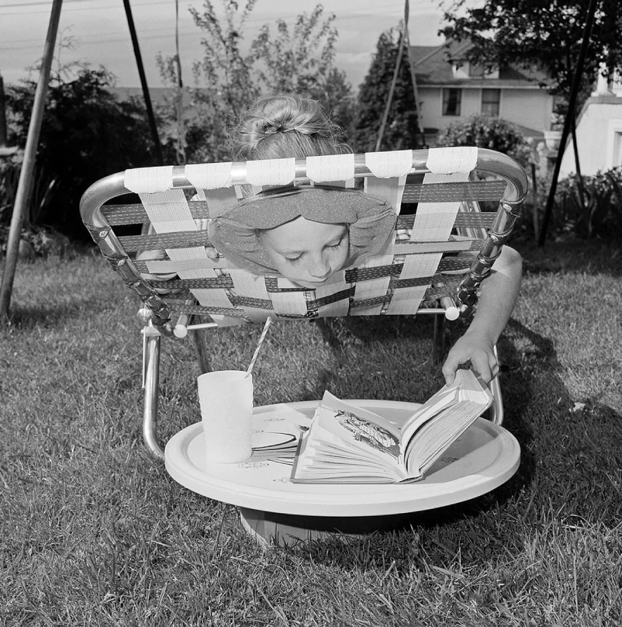 A person lies face-down on a lawn chair with their head poking through the woven straps, reading a book on a low table. A cup and another book sit nearby on the grass in a backyard setting.