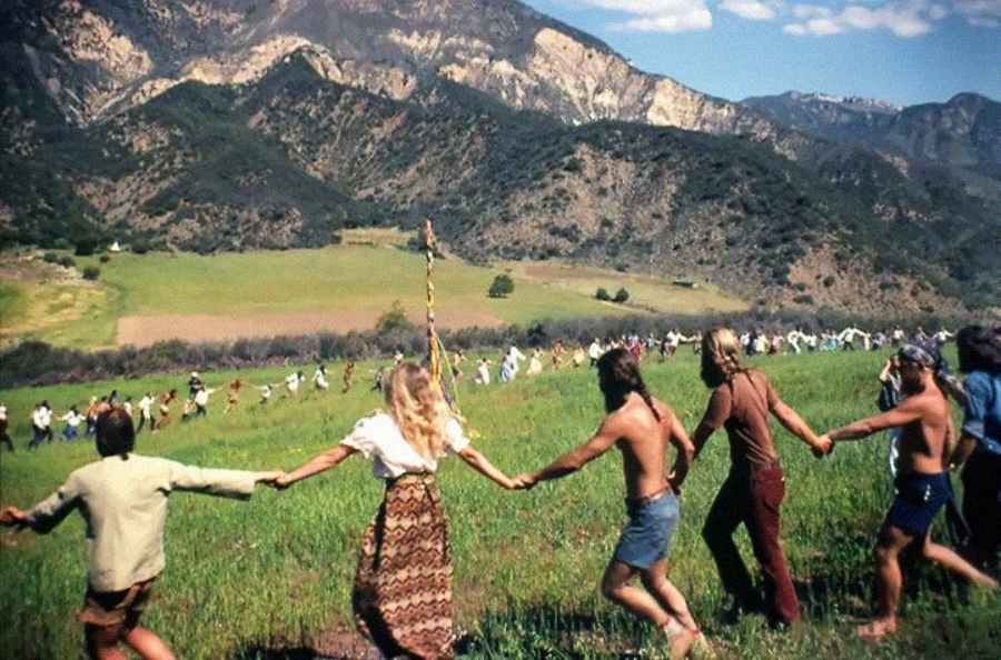 A large group of people holding hands in a circle on a grassy field, surrounded by mountains, under a partly cloudy sky. Some participants are barefoot and dressed in casual, bohemian clothing.