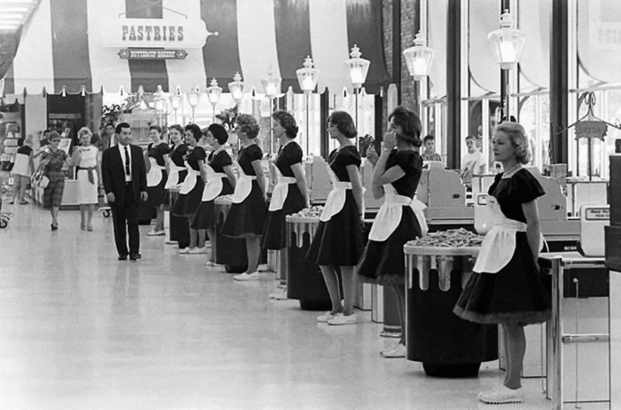 Women in matching uniforms and aprons stand in a row behind checkout counters in a vintage supermarket, while a man in a suit walks by. The scene has a 1950s or 1960s aesthetic.
