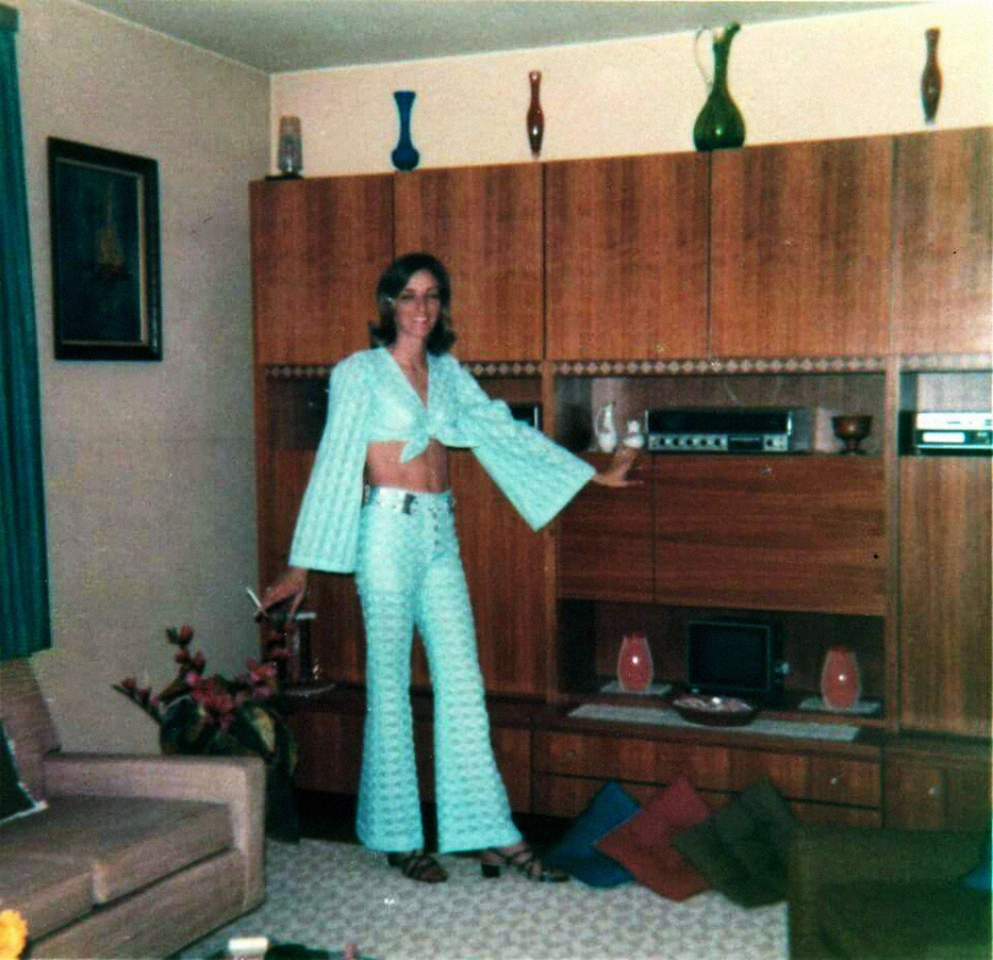 A person in a light blue patterned pantsuit stands in a retro living room, posing near a wooden wall unit with vases and electronics. Colorful pillows are on the floor and a drink is held in one hand.